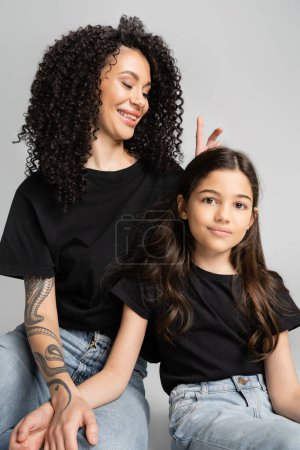 Foto de Sonriente madre sosteniendo los dedos cerca de la cabeza de la hija como orejas de conejo sobre fondo gris - Imagen libre de derechos
