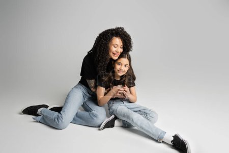 Smiling woman in t-shirt hugging preteen daughter while sitting on grey background 