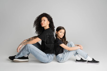 Tattooed mother and kid in jeans and t-shirts sitting back to back on grey background 