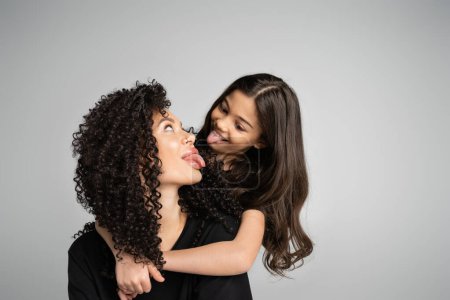 Preteen girl hugging mother and sticking out tongue isolated on grey 