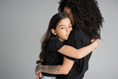 Preteen daughter in t-shirt hugging tattooed mom isolated on grey 