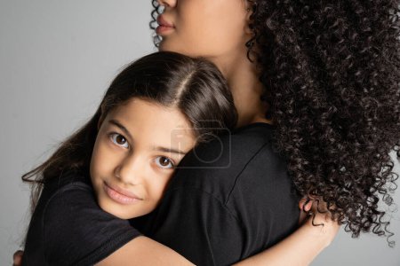 Portrait of preteen child hugging curly mom and looking at camera isolated on grey 