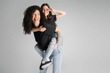 Positive and tattooed mother piggybacking preteen daughter showing peace sign isolated on grey 
