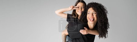 Positive and tattooed mother piggybacking preteen daughter showing peace sign isolated on grey, banner 