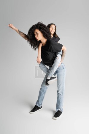 tattooed mother pouting lips and piggybacking preteen daughter on grey background 