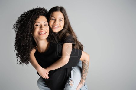 Positive Frau hält Tochter und blickt in Kamera isoliert auf grau 