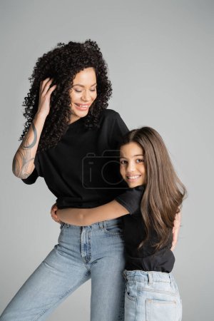 Smiling preteen girl hugging tattooed mom in t-shirt isolated on grey 