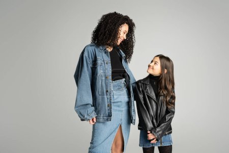 Cheerful woman in denim jacket looking at daughter isolated on grey 