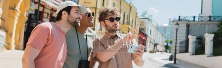 jeune guide en lunettes de soleil tenant smartphone et pointant du doigt près des touristes multiethniques sur la descente Andrews à Kiev, bannière