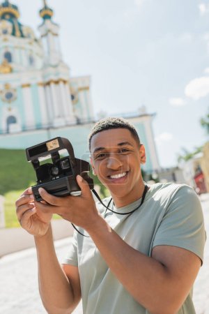 carefree african american tourist with vintage camera smiling near blurred St Andrews Church in Kyiv