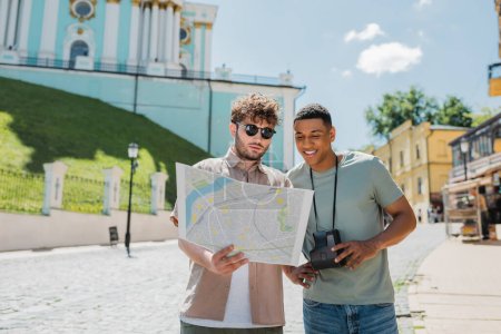Reiseleiter mit Sonnenbrille und afrikanisch-amerikanischer Reisender mit Oldtimer-Kamera beim Blick auf den Stadtplan auf der Andrews-Abfahrt in Kiew, Ukraine