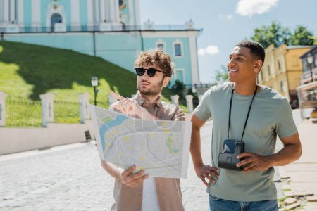 Reiseleiter hält Karte in der Hand und zeigt mit dem Finger in die Nähe eines lächelnden afrikanisch-amerikanischen Touristen mit Vintage-Kamera auf der Andrews-Abfahrt in Kiew