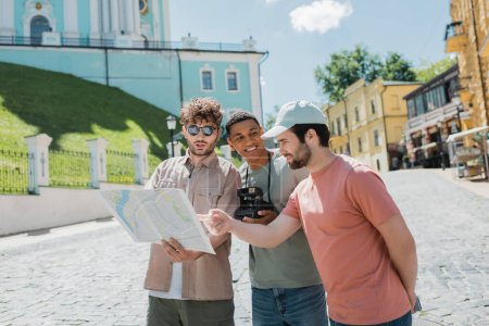 Foto de Turista barbudo apuntando al mapa de la ciudad cerca de guía turístico y hombre afroamericano sonriente con cámara vintage en el descenso de Andrews en Kiev - Imagen libre de derechos