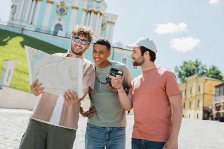 Lächelnder Reiseleiter mit Sonnenbrille hält Stadtplan in der Nähe multiethnischer Touristen mit Vintage-Kamera auf der Andrews-Abfahrt in Kiew