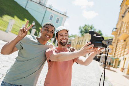 sonriente hombre afroamericano mostrando un signo de paz cerca de un amigo barbudo tomando selfie en una cámara vintage en el descenso de Andrews en Kiev