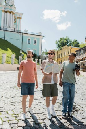 full length of interracial tourists with vintage camera and city map walking with guide on Andrews descent in Kyiv