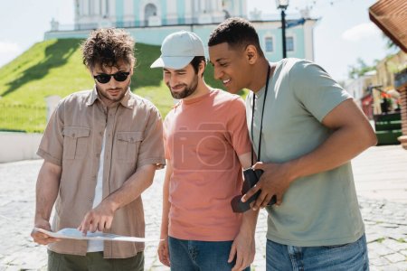 Reiseleiter mit Sonnenbrille zeigt auf die Reisekarte in der Nähe lächelnder multiethnischer Touristen beim Andreasabstieg in Kiew