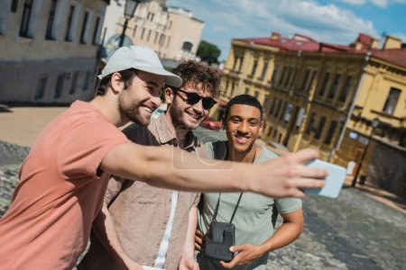 happy tourist in sun cap taking selfie with multiethnic men during excursion on Andrews descent in Kyiv