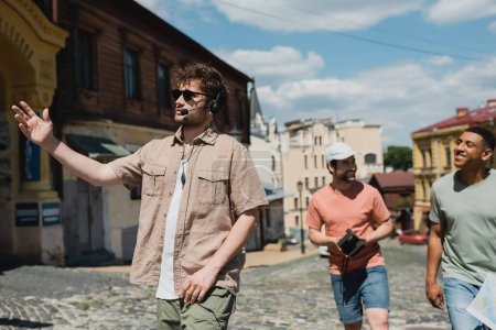 joven guía en auriculares y gafas de sol señalando con la mano durante la excursión con hombres interracial en el distrito de Podil en Kiev