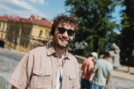 portrait of curly and carefree tour guide in sunglasses and headset smiling near blurred multiethnic travelers on Andrews descent in Kyiv