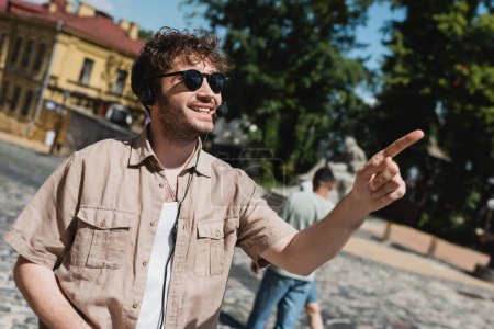fröhlicher Reiseleiter mit Headset und Sonnenbrille, der mit dem Finger auf verschwommene Abfahrten in Kiew zeigt