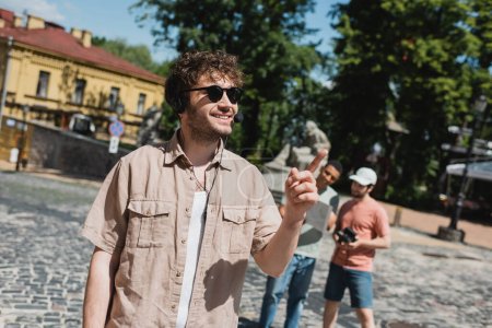Reiseleiter mit Sonnenbrille und Headset lächelt und zeigt mit dem Finger auf verschwommene multiethnische Touristen auf der Andreasabfahrt in Kiew