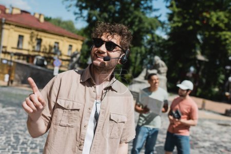 guía joven y despreocupado en gafas de sol y auriculares apuntando con el dedo cerca de los turistas interracial borrosa en el distrito de Podil en Kiev