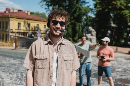 carefree tour guide in headset and sunglasses smiling near blurred multiethnic travelers on Andrews descent in Kyiv