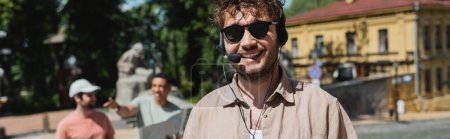 retrato de guía turístico joven en auriculares y gafas de sol cerca de los turistas interracial borrosa en el descenso de Andrews en Kiev, bandera