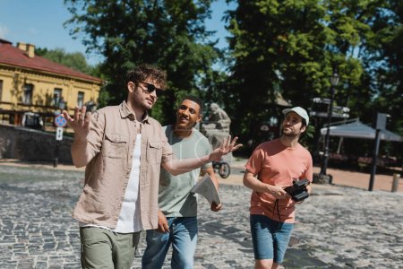 Reiseleiter mit Sonnenbrille gestikuliert, während er mit multikulturellen Touristen während eines Spaziergangs auf der Andreasabfahrt in Kiew spricht