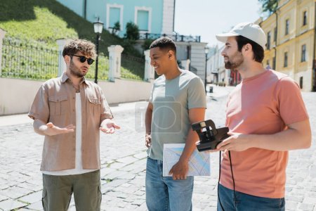 junger Reiseleiter mit Sonnenbrille spricht auf der Andreasabfahrt in Kiew mit Vintage-Kamera und Landkarte zu interrassischen Männern