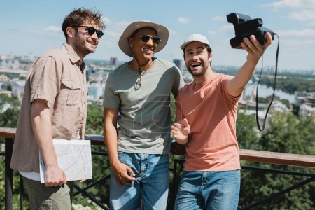 Unbekümmerter bärtiger Mann mit Oldtimer-Kamera macht Selfie mit afrikanisch-amerikanischem Freund und Reiseleiter in der Nähe von Zaun im Stadtpark