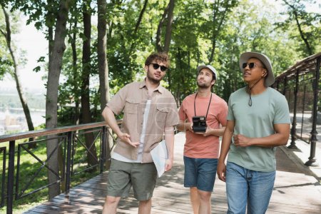 viajeros multiculturales en sombreros mirando hacia otro lado durante el paseo de verano con guía turístico en el parque urbano