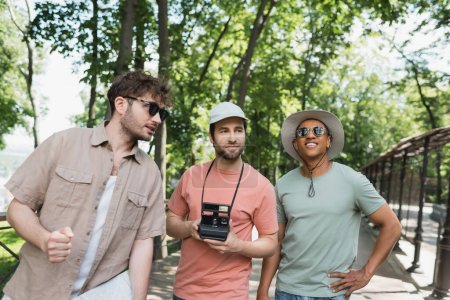 joven guía de turismo en gafas de sol hablando con turistas multiétnicos en sombreros durante la excursión en el parque urbano