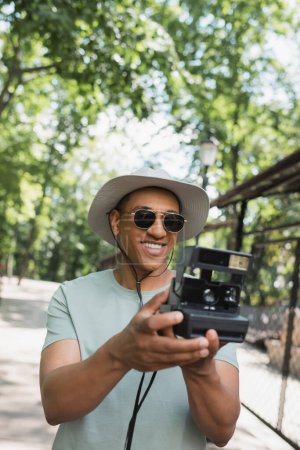 smiling african american traveler in sun hat and sunglasses taking photo on vintage camera in summer park