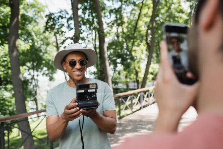 verschwommener Mann mit Smartphone fotografiert unbeschwerten afrikanisch-amerikanischen Touristen in Sonnenbrille posiert mit Vintage-Kamera im Park
