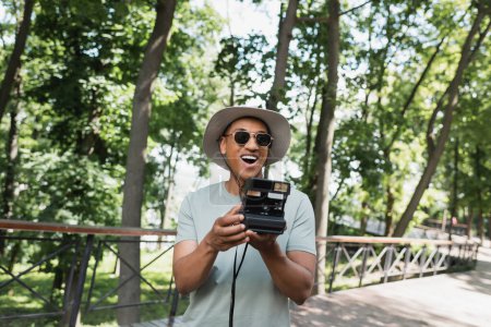 Téléchargez les photos : Stupéfait voyageur afro-américain portant des lunettes de soleil et chapeau de soleil tout en prenant des photos sur la caméra vintage sur la passerelle dans le parc - en image libre de droit