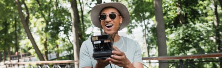 Téléchargez les photos : Excité voyageur afro-américain en chapeau de soleil et lunettes de soleil prenant des photos sur caméra vintage dans le parc d'été, bannière - en image libre de droit