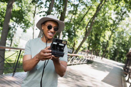 Téléchargez les photos : Heureux touriste afro-américain en chapeau de soleil et lunettes de soleil prenant des photos sur caméra vintage sur passerelle dans le parc de la ville - en image libre de droit