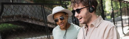 joven guía turístico en gafas de sol y auriculares sonriendo cerca de turista afroamericano en sombrero de sol, bandera