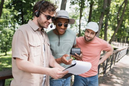 homme afro-américain joyeux avec caméra vintage pointant vers presse-papiers près du guide dans le casque pendant l'excursion dans le parc de la ville