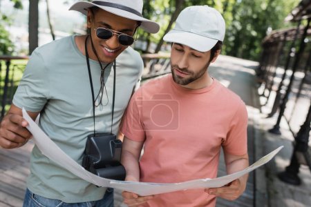 lächelnder afrikanisch-amerikanischer Reisender mit Oldtimer-Kamera beim Betrachten der Reisekarte mit Freund im Sommerpark