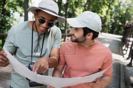 Lächelnder afrikanisch-amerikanischer Tourist mit Sonnenbrille zeigt auf Karte neben bärtigem Freund im Sommerpark