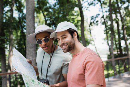 positive multiethnic tourists in sun hats looking at travel map in blurred park