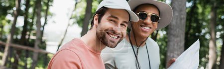 fröhliche Touristin blickt in die Kamera neben einem afrikanisch-amerikanischen Freund mit Sonnenbrille und hält Reisekarte im Park, Banner