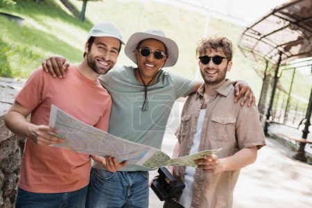 carefree african american man in sun hat and sunglasses embracing friends on urban street
