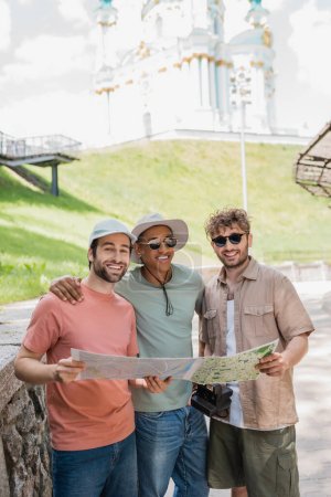 glückliche multiethnische Touristen mit Sonnenbrille, Stadtplan in der Hand und lächelnd in der Nähe der St. Andrews Kirche auf verschwommenem Hintergrund 