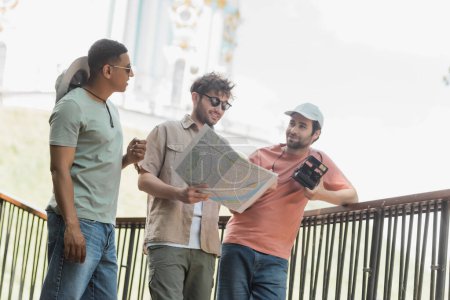 Photo for Smiling man in sunglasses looking at map near multiethnic friends with vintage camera on city street - Royalty Free Image