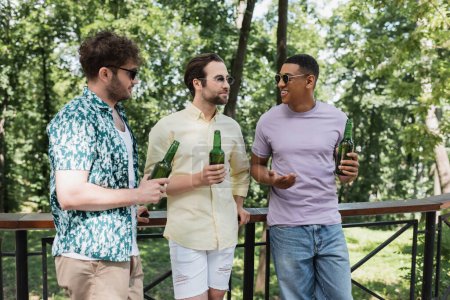 amigos multiétnicos felices y elegantes en gafas de sol con cerveza fresca y hablando cerca de valla en el parque de la ciudad