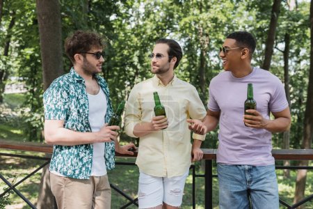 fröhlicher afrikanisch-amerikanischer Mann im Gespräch mit trendigen Freunden, die mit Bier am Zaun im Stadtpark stehen